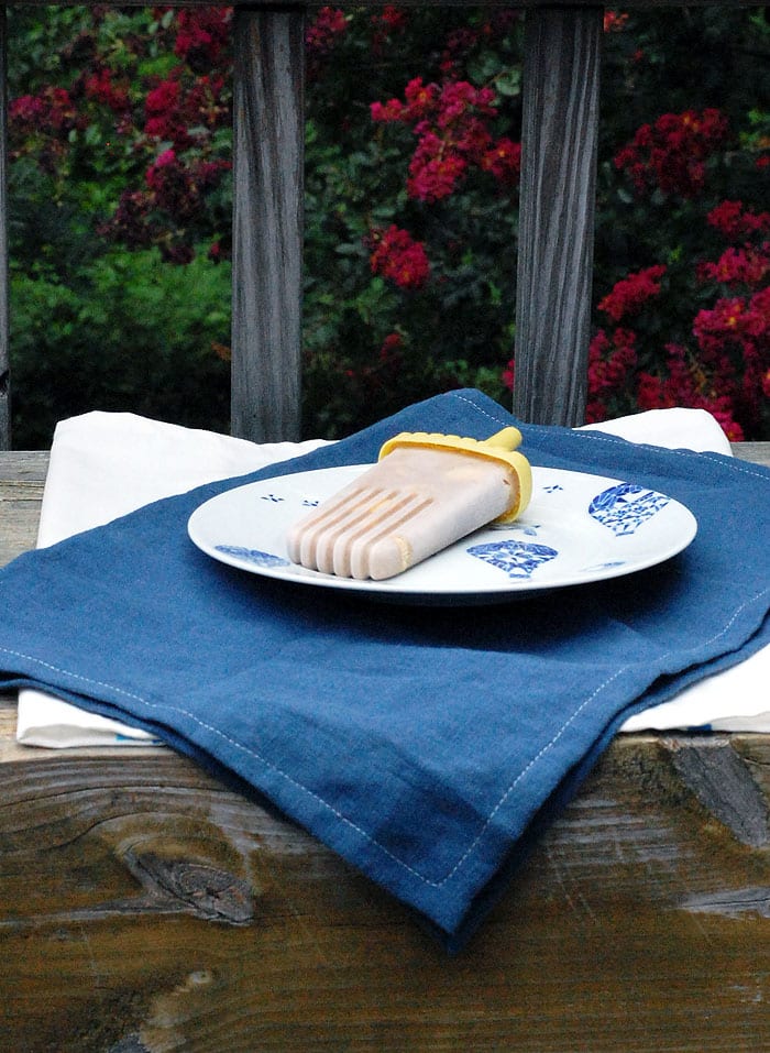a vegan banana pudding popsicle on a decorated white plate placed on top of a blue cloth and another white cloth on top of a wooden table with greens and red flowers on the background