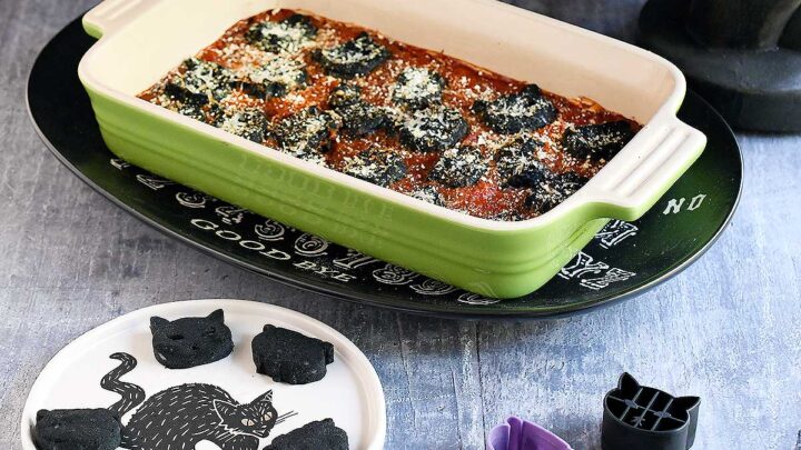 Cat and cauldron shaped black vegan gnocchi baked in a tomato sauce in a green casserole dish in a Halloween setting.